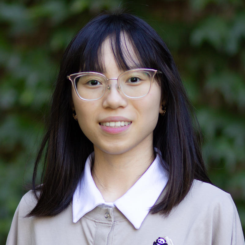 Headshot of young woman with shoulder length black hair, wearing glasses, smiling showing teeth, with blurred green background