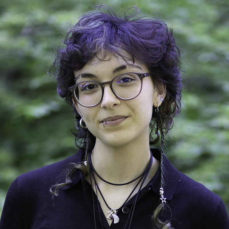 Headshot of young woman smiling with lips closed, with wavy, short dark hair, wearing glasses, multiple ear piercings, lip piercing, and necklaces, with blurred foliage in the background