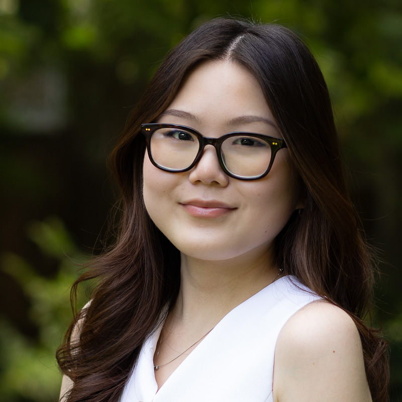 Headshot of Hannah Won, with long, dark, wavy hair, wearing glasses, white v-neck, sleeveless top, smiling not showing teeth