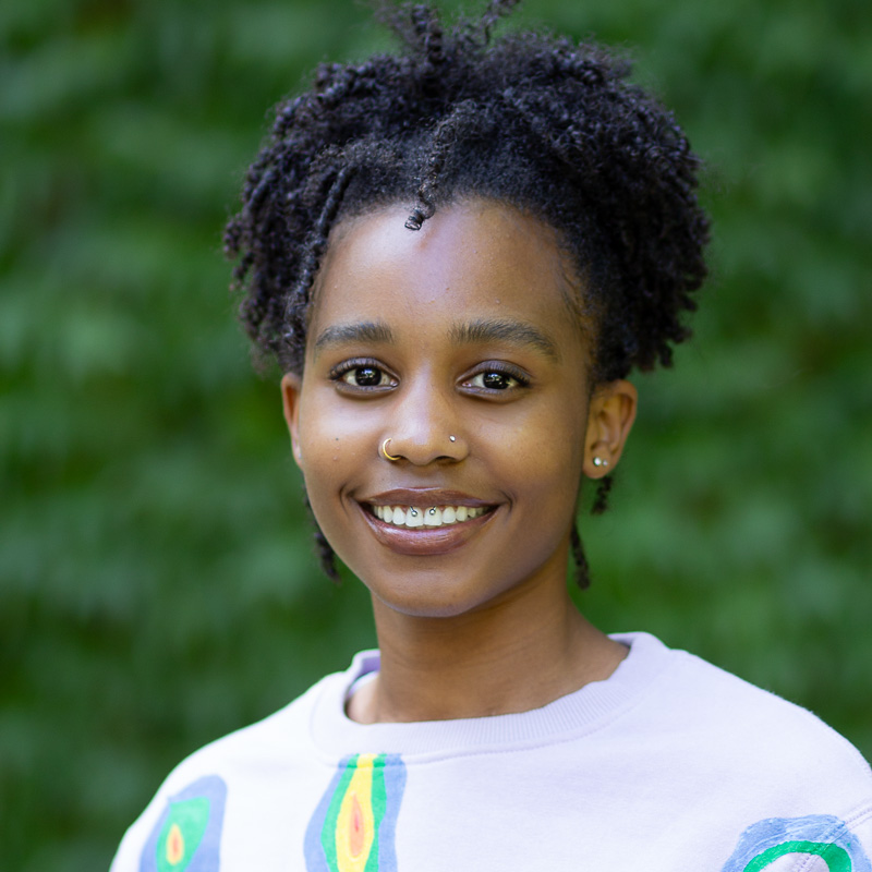 Headshot of Jeanne Bilap-Nsegbe with short dark curly hair, smiling showing teeth, wearing light top with colourful graphics, greenery in the background