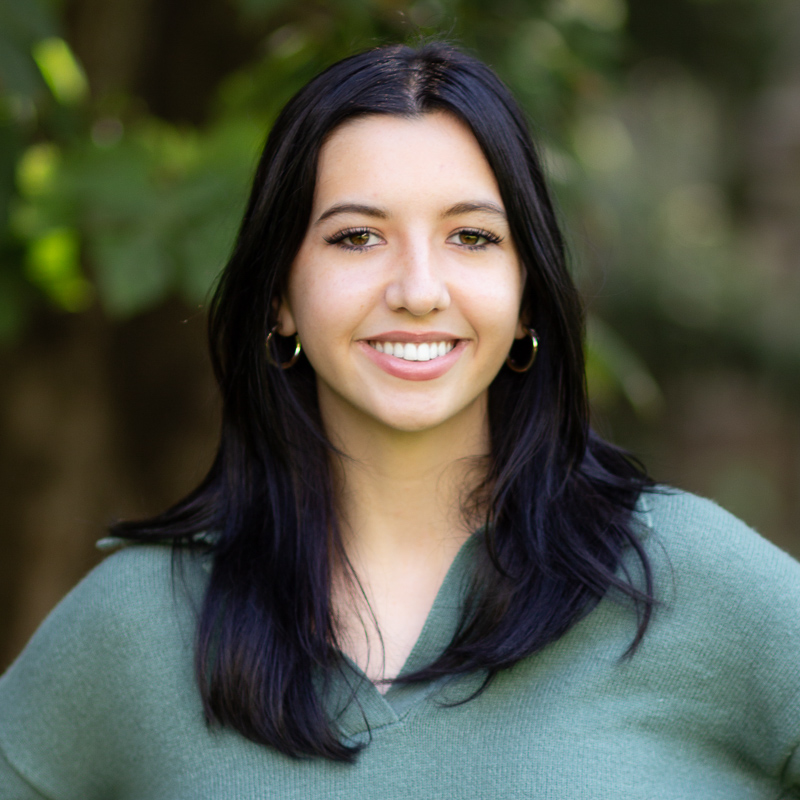headshot of Jordan, smiling showing teeth, with shoulder-length dark hair, wearing green v-neck sweater