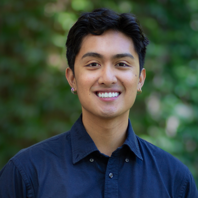 Headshot of young man with short, black hair, smiling showing teeth, small hoop earrings, blue shirt, with green blurred background