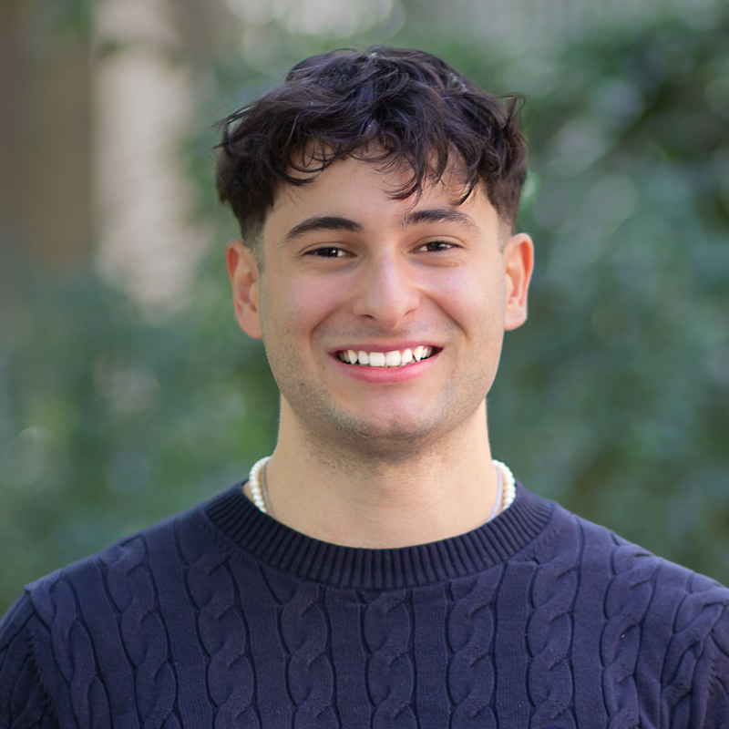 Headshot of Lucas Schiavello, with short, dark wavy hair, smiling showing teeth, wearing navy sweater