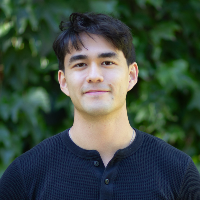 Headshot of young man  with short dark hair, smiling without showing teeth, wearing nay shirt, against blurred green background
