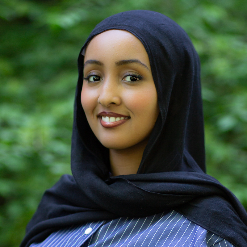 Headshot of young women with medium brown skin, smiling showing teeth, wearing navy headscarf, blue shirt with white stripes, with green foliage blurred in background