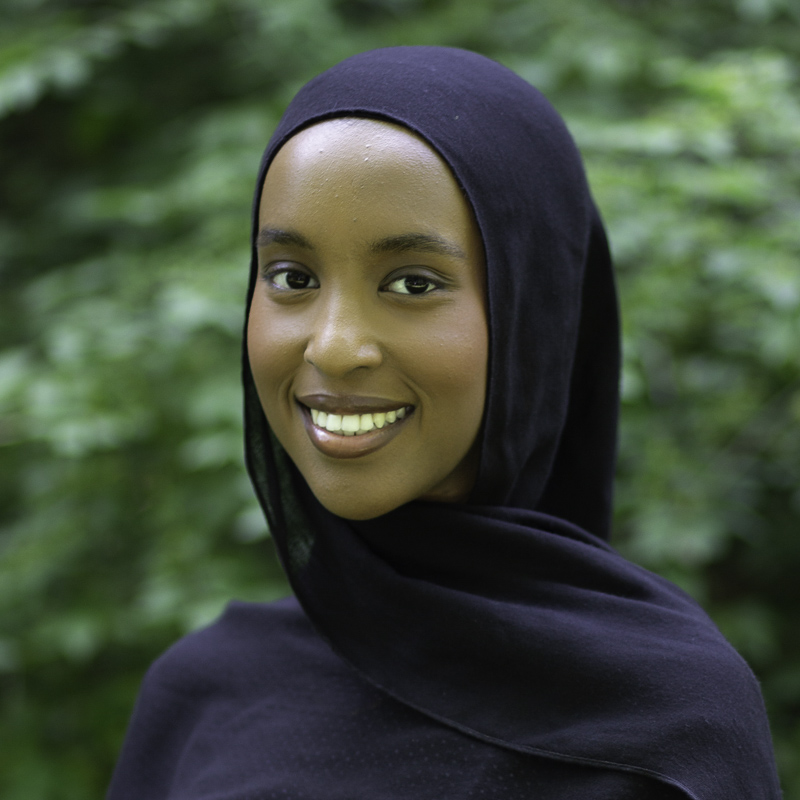 Headshot of young women with medium brown skin, smiling showing teeth, wearing navy headscarf, with green foliage blurred in background