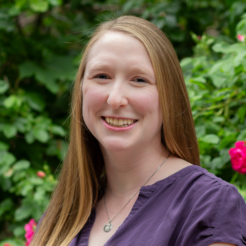 Headshot of Leigh Paulseth with long blonde hair, smiling showing teeth, wearing purple shirt, with greenery in the background