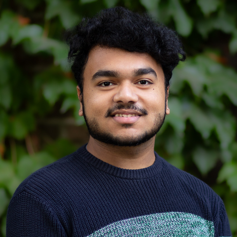 Headshot of Vivek Dev Bishal with dark hair and beardm wearing navy sweater, smiling showing teeth, with greenery in the background