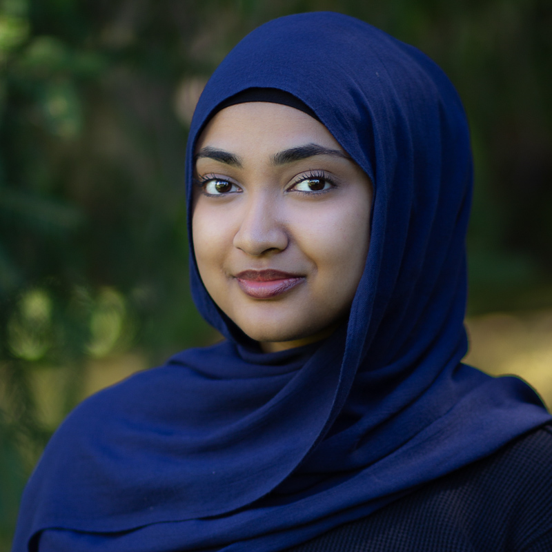Headshot of Yusra Saifuddin, wearing a navy headscarf, smiling gently