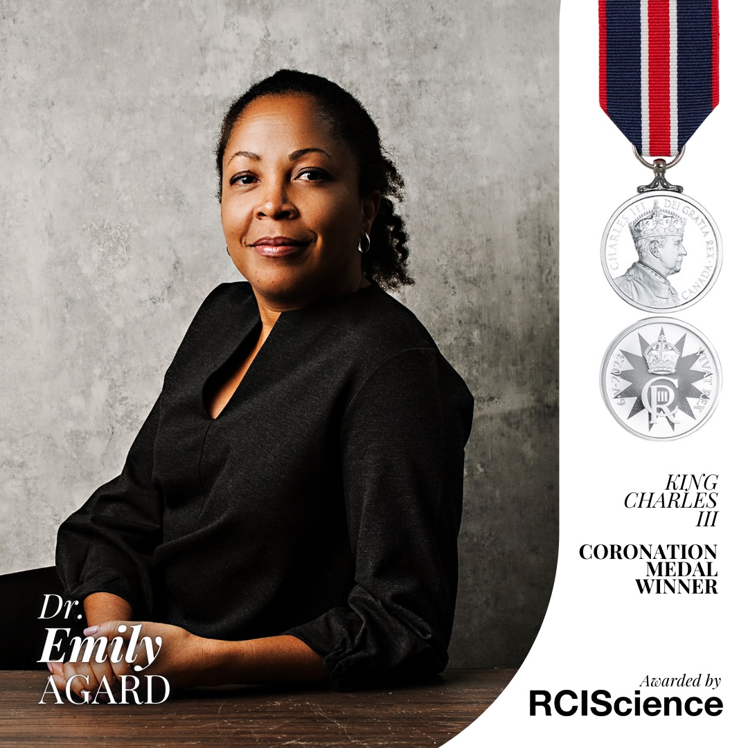 Emily agard smiling, wearing black top, seated, leaning on table, smiling with mouth closed and images of coronation medal on the right side