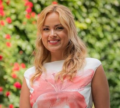 Kayla Bourdeau, with wavy blonde hair, smiling showing teeth, wearing white and lforal sleevelss top against blurred greenery