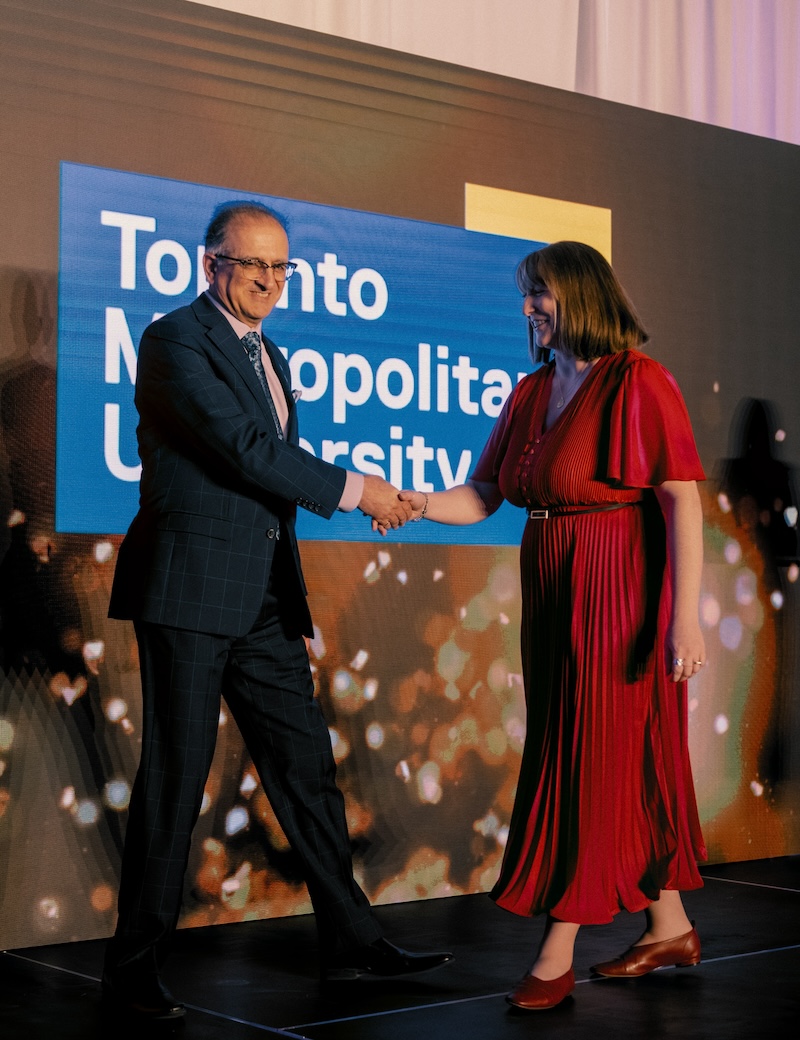 Leigh Paulseth accepting award on stage, wearing long red dress, red shoes, shaking the hand of presenter, with toronto metropolitan University logo on the wall in the background