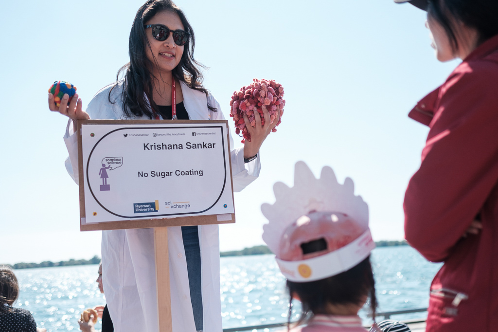 Soapbox speaker Krishana Sankar is standing on her soapbox holding props with her hands and engaging with participants.