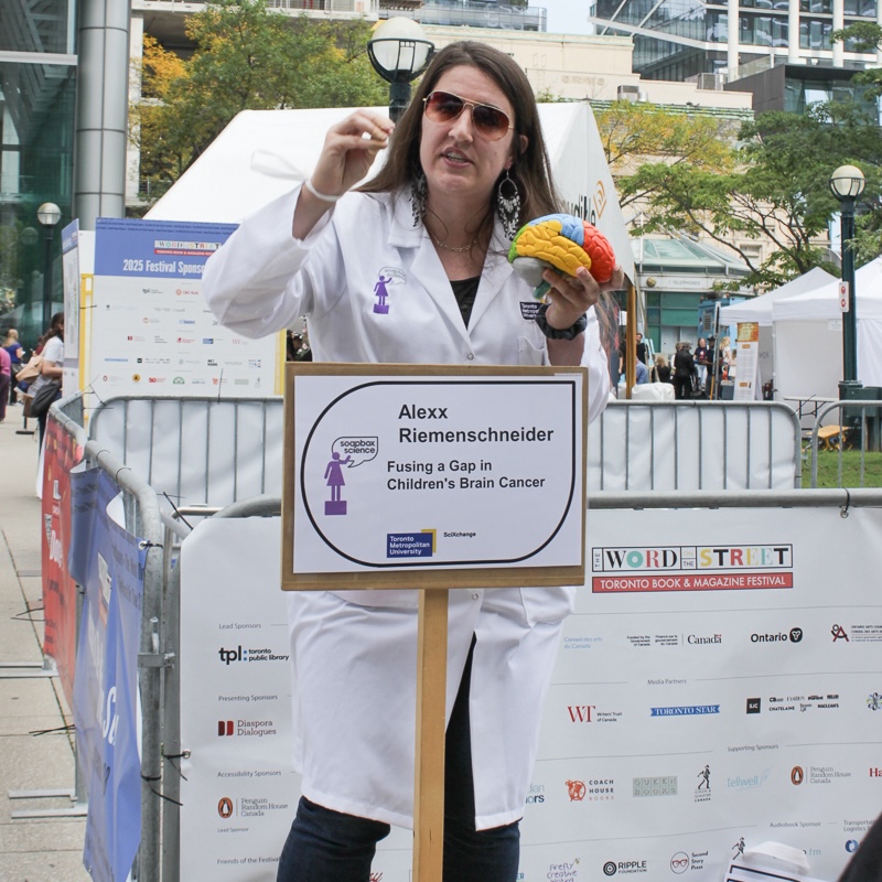 Young woman with dark hair, sunglasses, lab coat standing behind sign with her name and speaking topic  with city square in the background