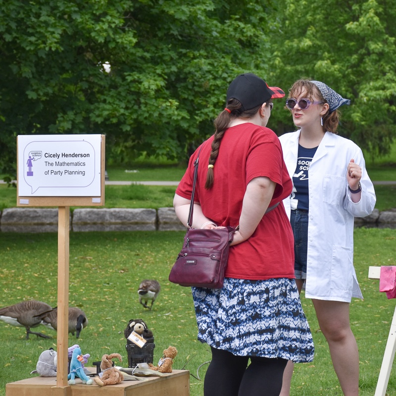 Young woman wearing sunglasses, black t-shirt, lab coat talking to woman in red shirt, with sign besid them and greenery in the background