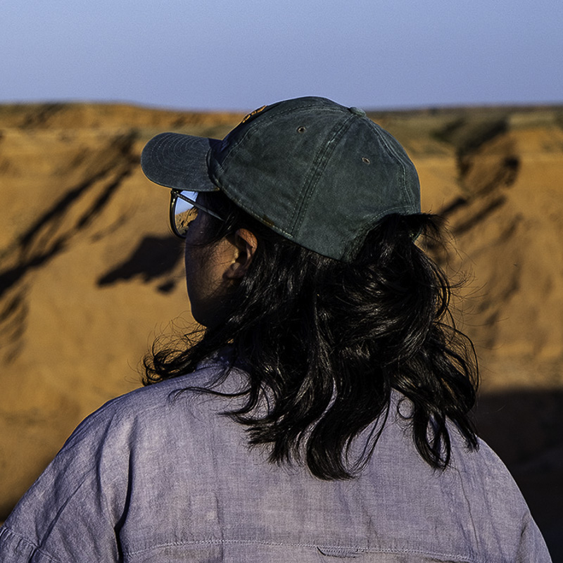 headshot of young woman with shoulder length dark hair, green baseball cap, glasses, facing away from the camera looking at an angle at golden rock iin the backgroundfacing away from 