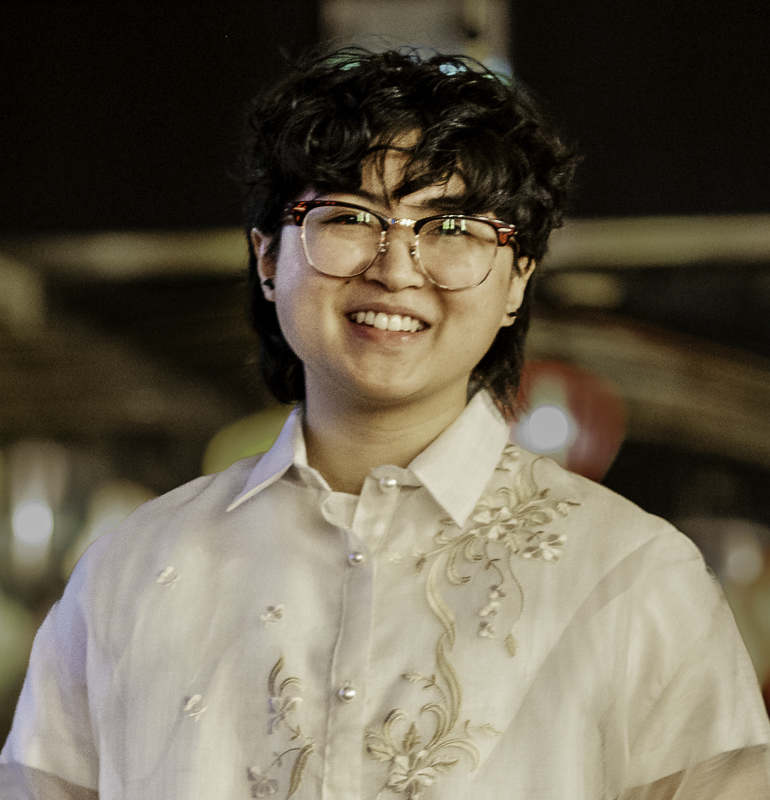 Headsot of Deny Maranga with short, wavy dark hair, glasses, smiling showing teeth, wearing beige shirt with embossed design,