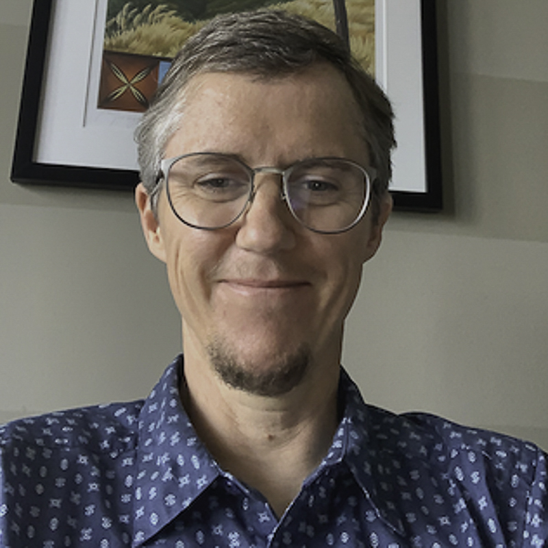 headshot of A W Peet wearing glasses and blue patterned shirt with beige wall and framed art in the background