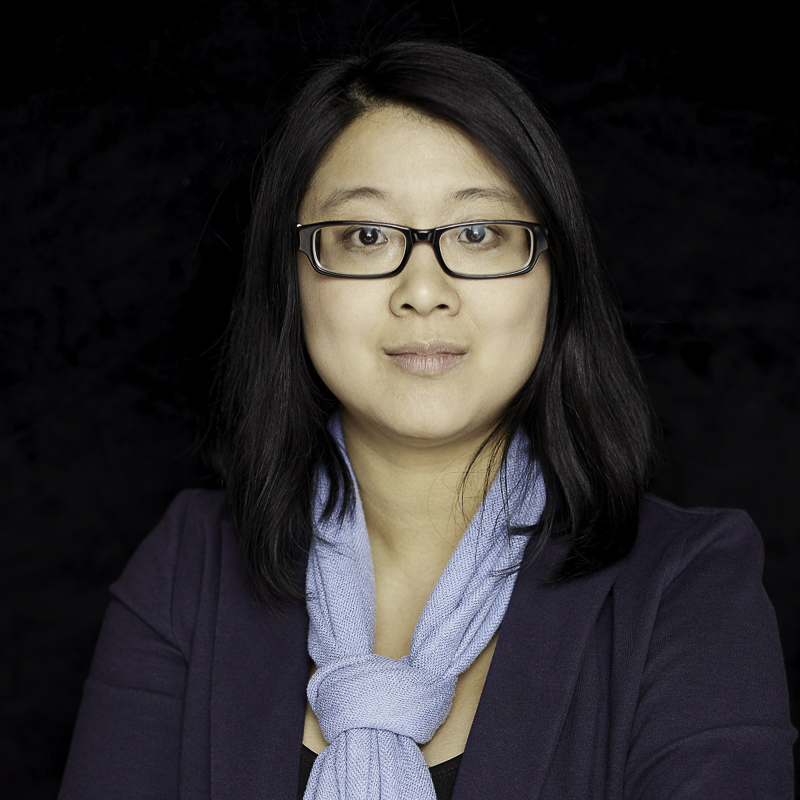 head shot of young woman with shoulder length black hair, wearing glasses, blue blzer and light blue scarf against a black background