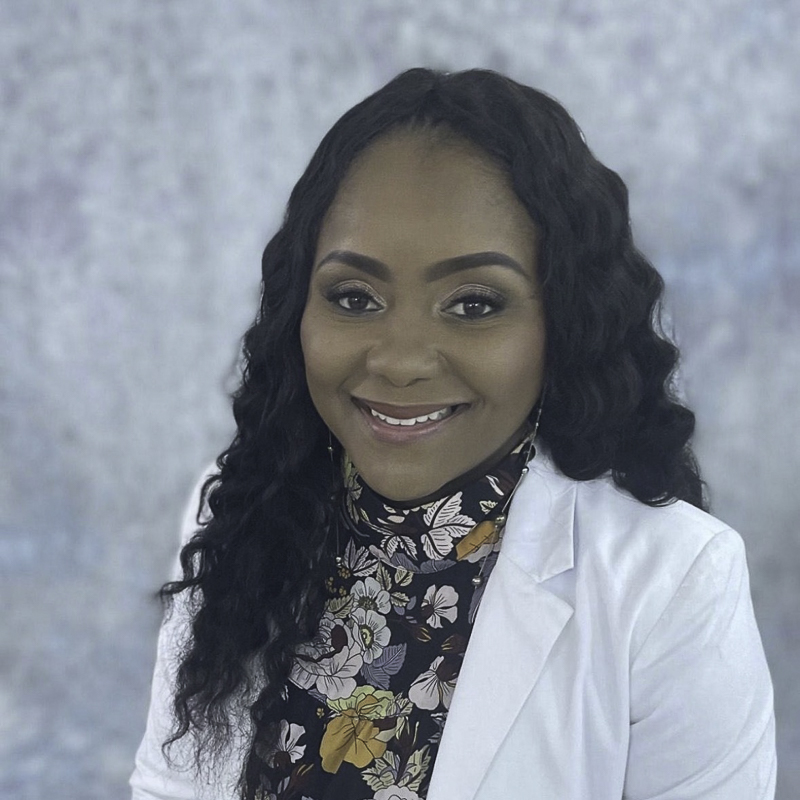 headshot of young woman with medium brown skin, black wavy hair, wearing white jacket and floral scarf