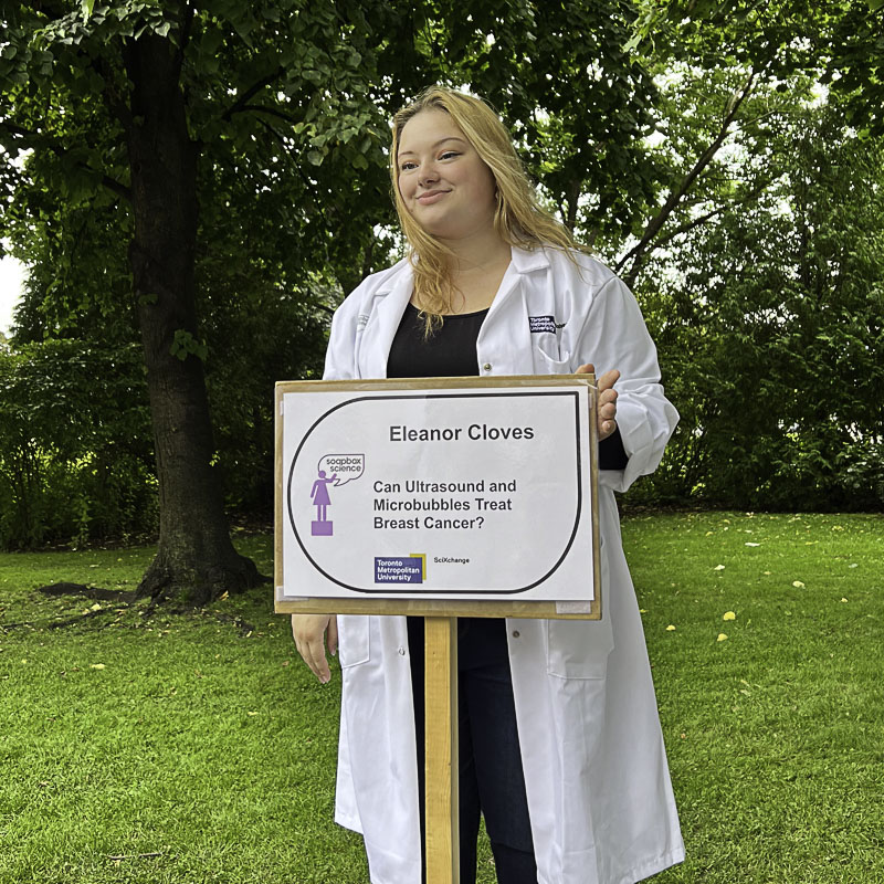 Young blonde woman wearing lab coat stands behind sign with her name and topic, greenery in background