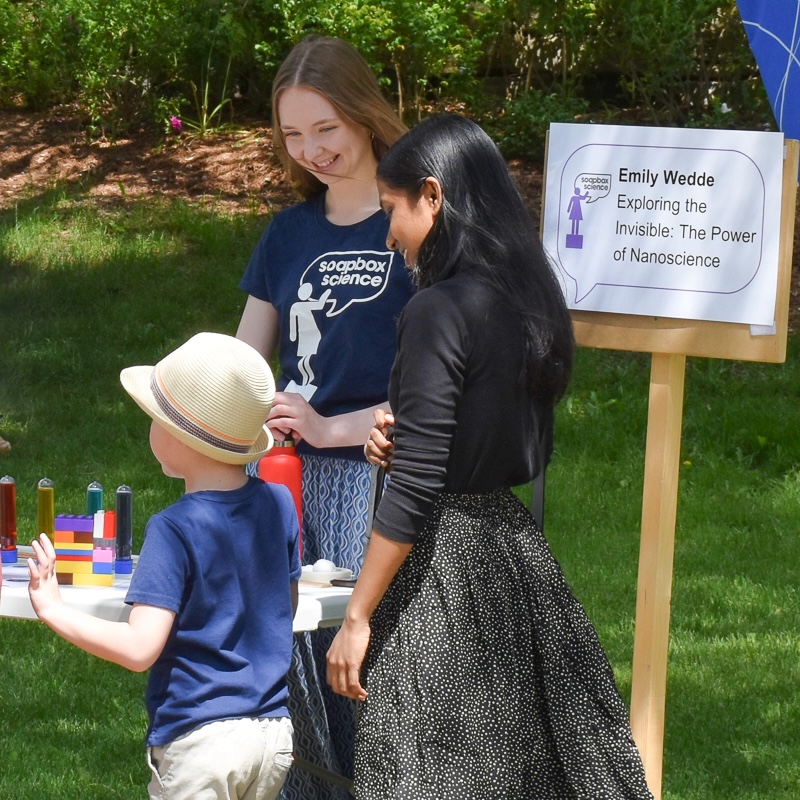 Young woman wearing t-shirt saying soapbox science stands talking to young boy and young woman with sign beside them and greenery in the background
