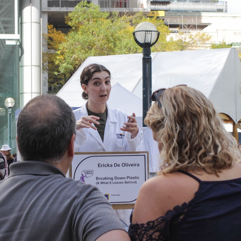 Young woman with brown hair, lab coat standing behind sign with her name and speaking topic, talking to man and woman with city square in background