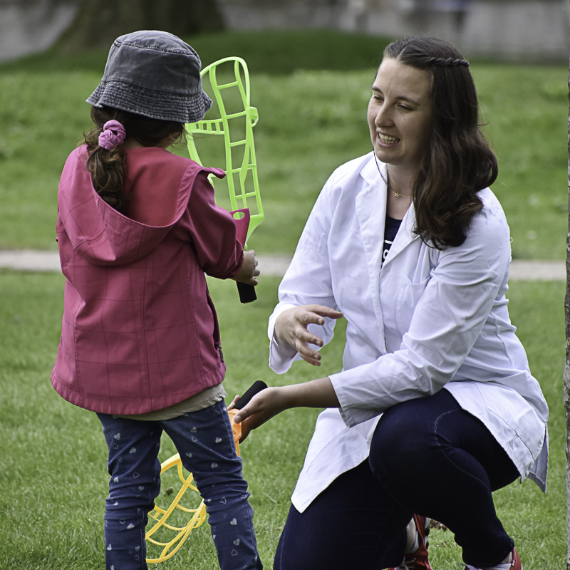 Young woman with brown hair, lab coat kneeling to talk to young girl wearing pink jacket, interacting with handheld props