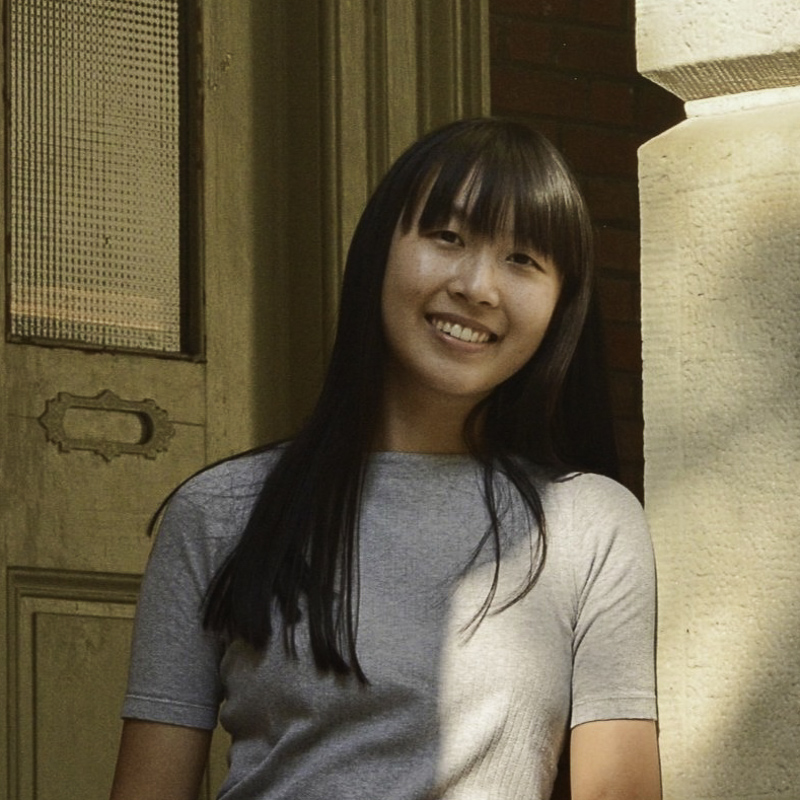 Young woman with long straight black hair with bangs, smiling showing teeth, leaning against a wall with a mix of window light and shadow against her face and body