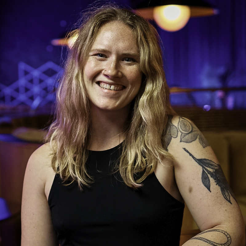 young woman with shoulder lenght blonde wavy hair, smiling, wearing black tank top, displaying tatoes on left shoulder and upper arm