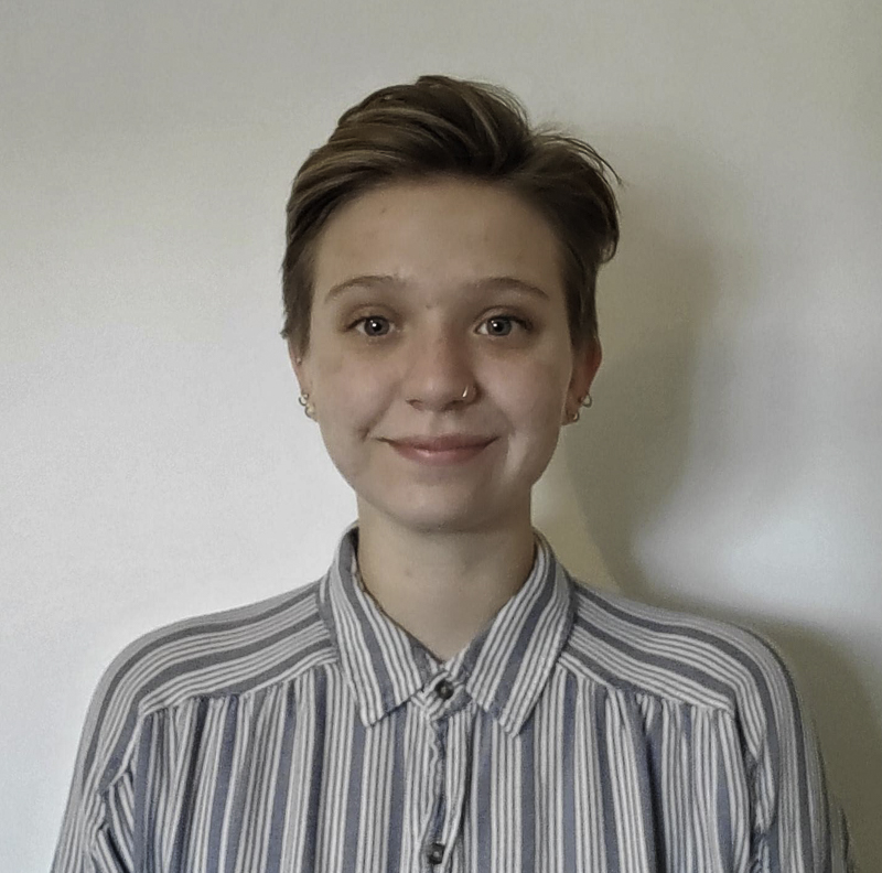 headshot of Mads Rose, with short light brown hair, small earrings, smiling, wearing striped shirt against a light background
