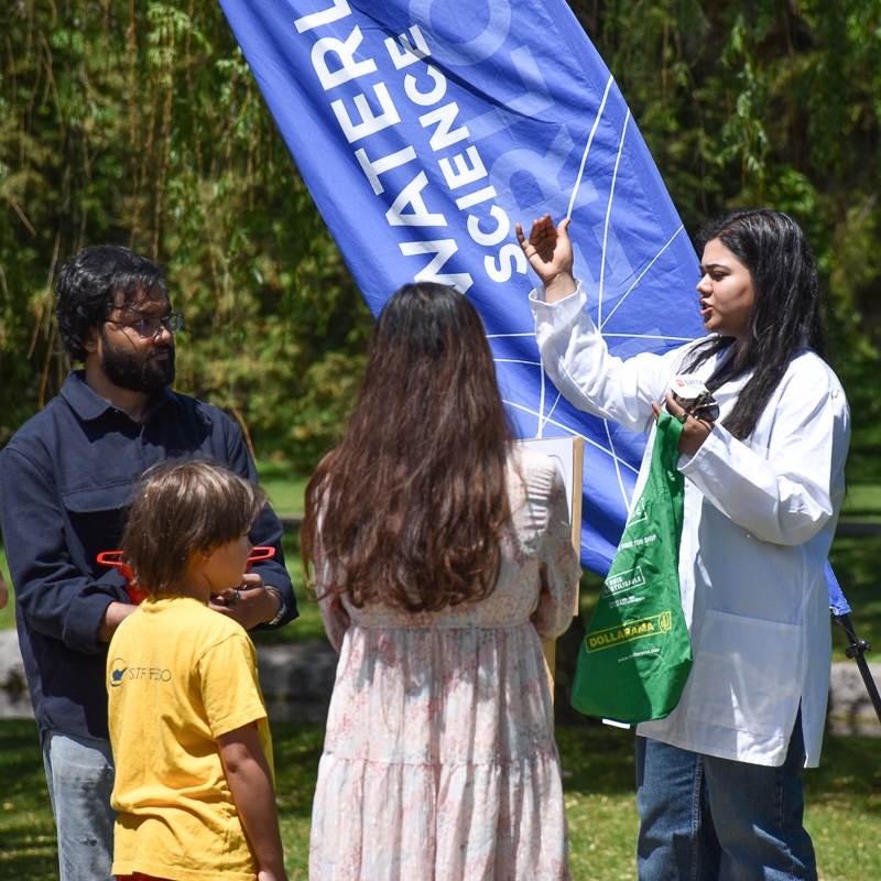 Young woman with long dark hair, wearing labcoat, holding green bag, talking to  a man, woman and child, with Waterloo science sign and greenery in the background