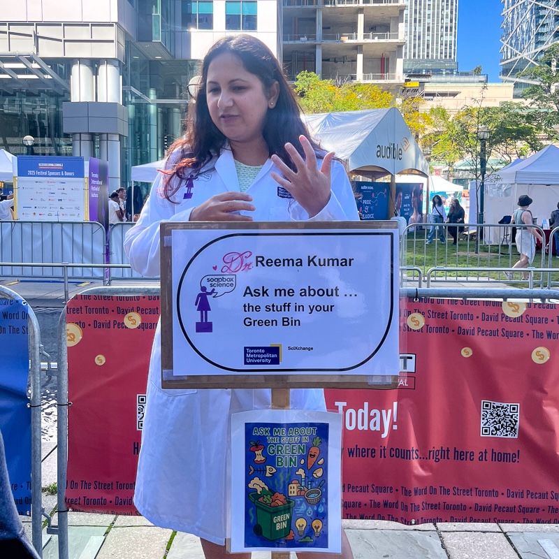 Young woman with dark hair, standing behind sign with her name and speaking topic,  with city square in the background