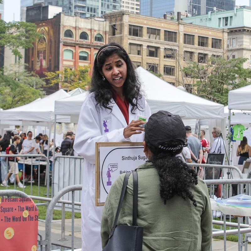 Young woman with dark hair, lab coat, standing behind sign with her name on it, talking to a woman, with city square and high-rise buildings in the background