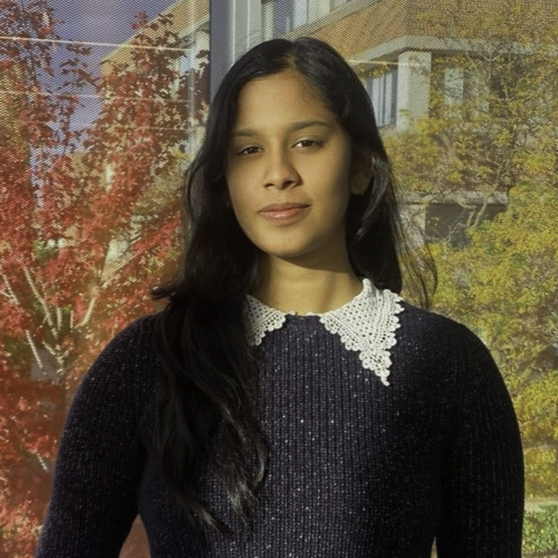 headshot of young woman with long black hair, smiling, wearing black knitted sweater with white knitted collar, with autumn foliage and a building in the background