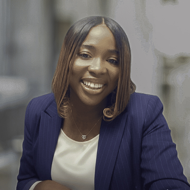Young woman with shoulder length straight black and brown hair, brown skin, smiling showing teeth, wearing navy pin-striped blazer over off-white top, background blurred