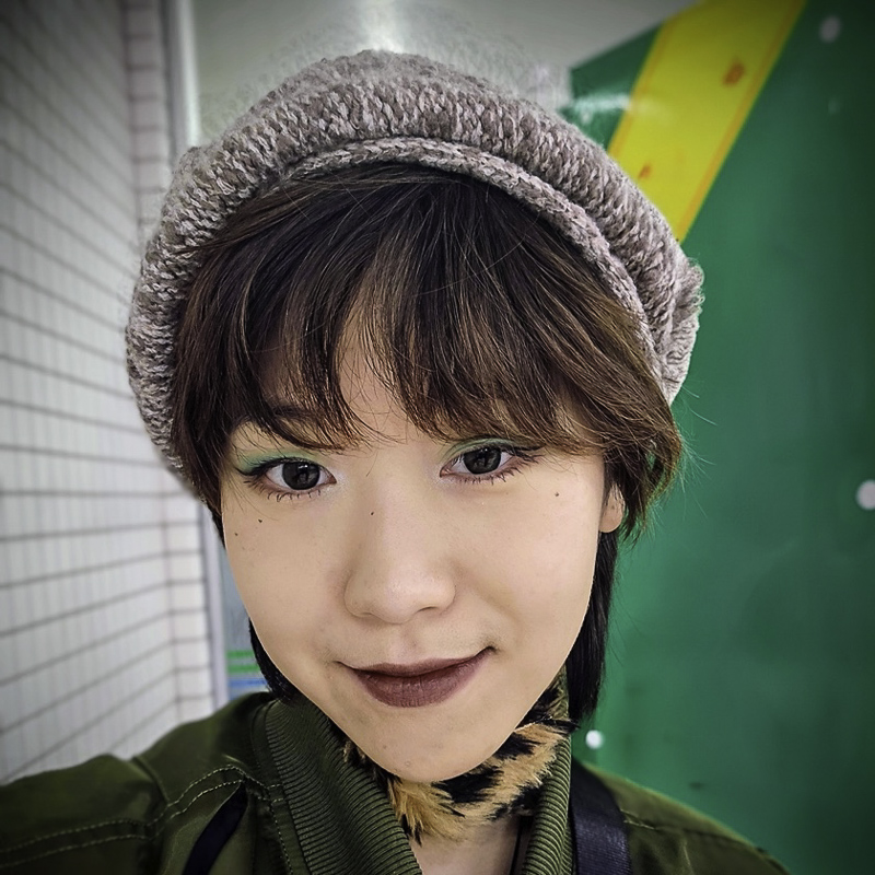 headshot of young woman wearing beigh knit cap over short dark hair, with bangs, green eyeshadow, smiling softly, wearing green top