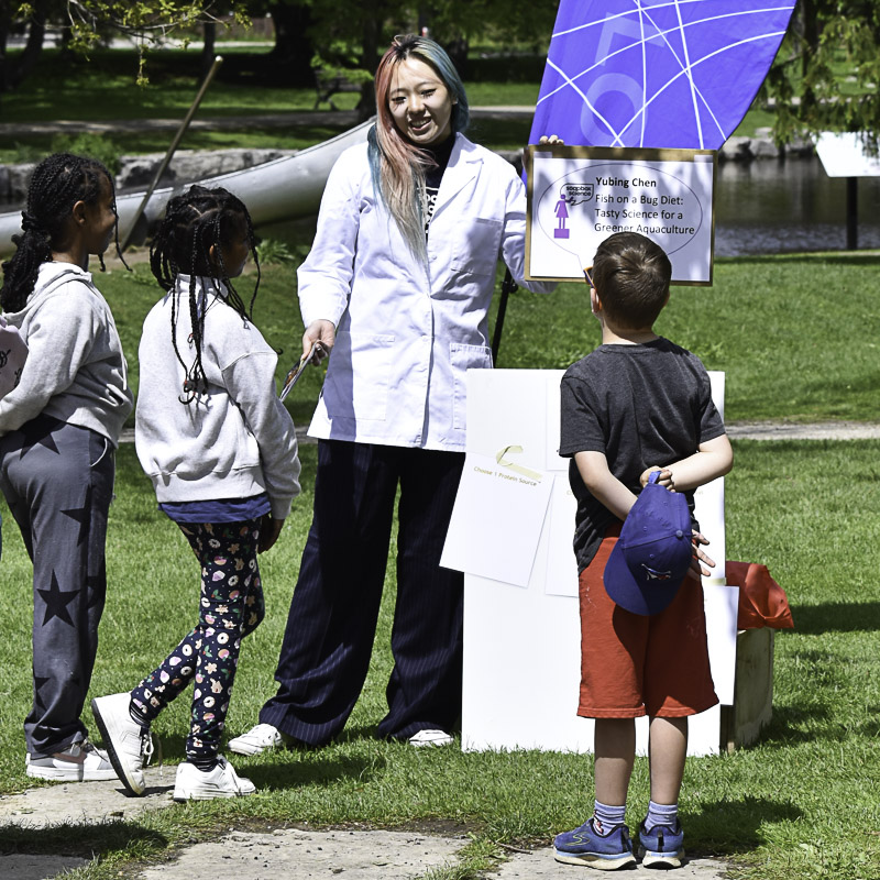 Young woman with hair dyed different colours, wearing lab coat, talking to 2 girls and a boy, with greenery in the background