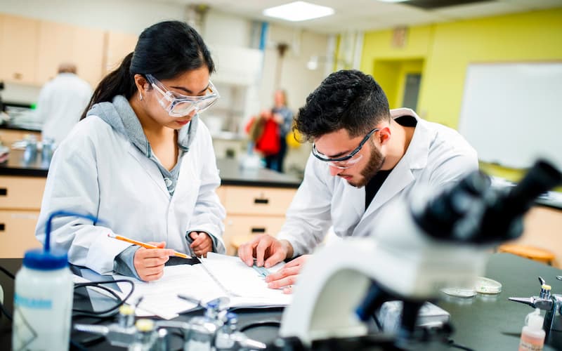 Two students wearing lab goggles and lab coats engrossed in a project, surrounded by scientific equipment and materials.
