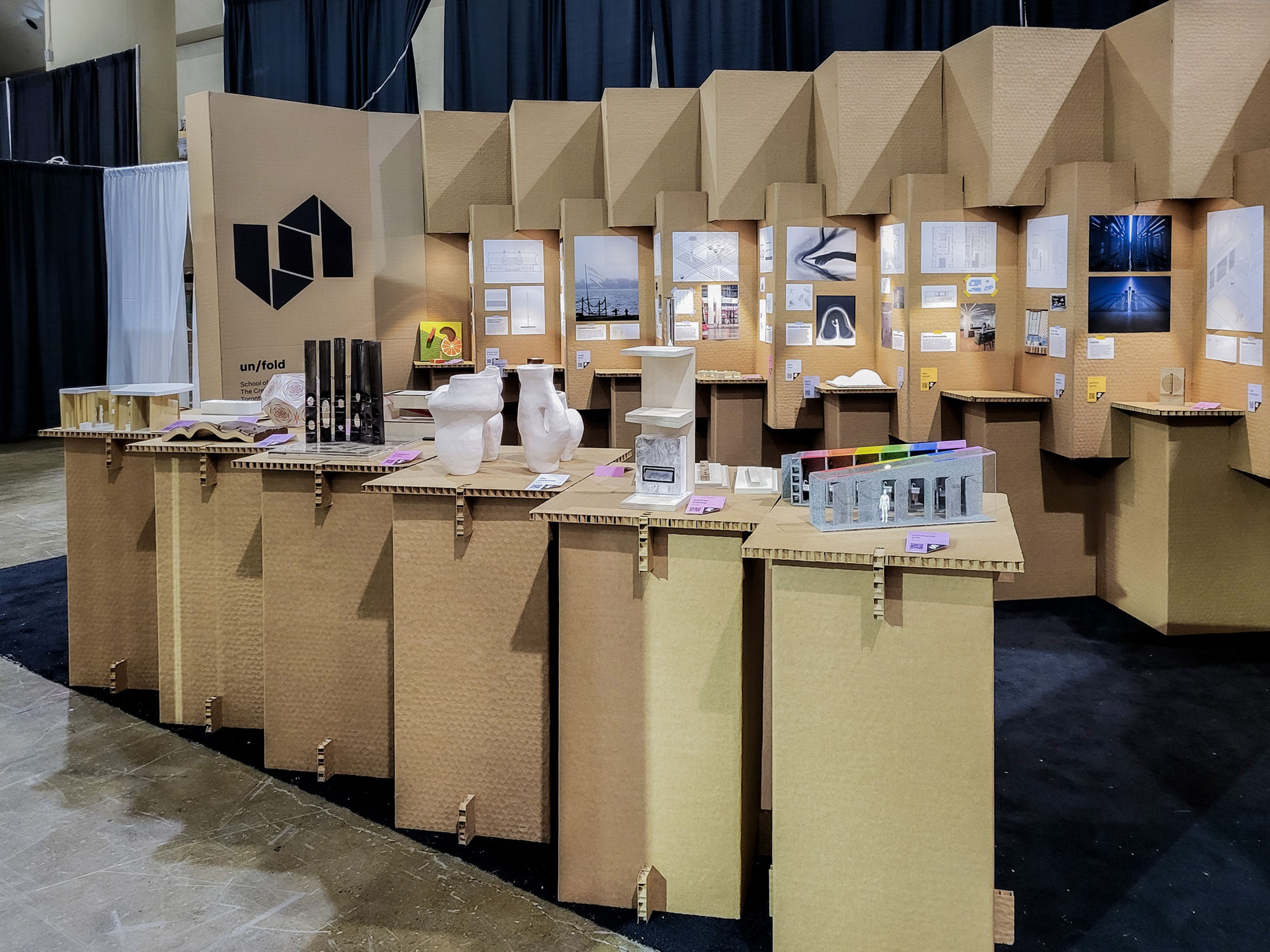  A brown cardboard booth on a black carpet with plinths in front of it. The booth unfolds and folds, revealing student work in every corner and fold.