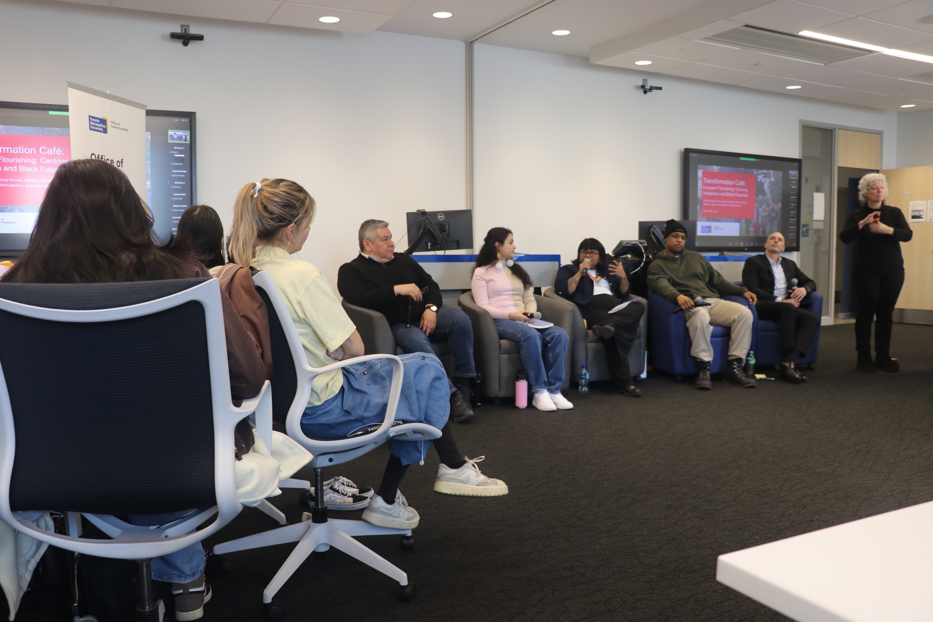 Photo of Transformation Cafe - Insurgent Flourishing: Centring Indigenous and Black Futurities panelists. Attendees look on. ASL interpreter to the side. 