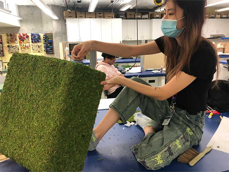 A woman wearing a blue surgical mask with long hair sits cross-legged on a table, putting together a wooden box covered in fake grass.
