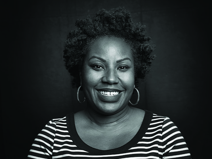 A black and white photo of a woman with short curly hair sitting in front of a black wall. The woman is wearing hopped earrings and a striped shirt.