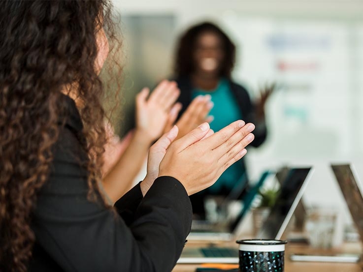 People sitting at desk watching a presentation and clapping. Woman presenting blurred in the background.