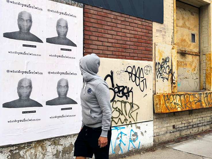 Man in grey hooded sweatshirt walking past a brick wall with black and white posters.