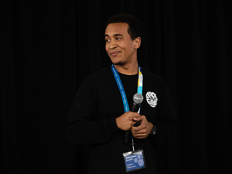 Sheldomar Elliott holding microphone in hand, looking to the left when smiling, standing in front of black backdrop. 