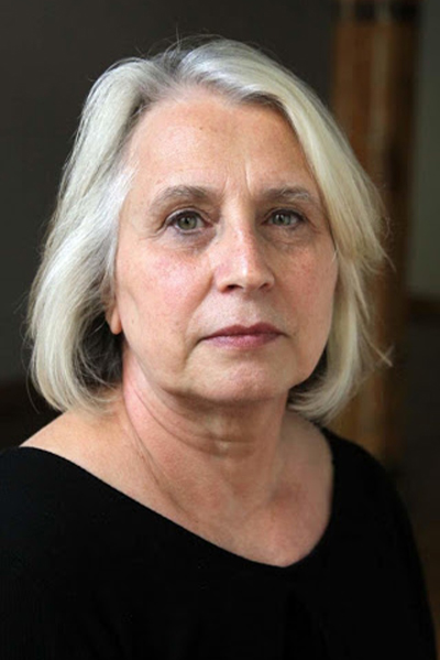 A woman with short white hair sitting in front of a back background. She is wearing a black shirt.