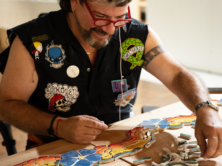 Man in a black cut-off shirt covered with badges working with mosaic tiles  at a table. 