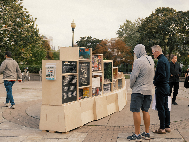 Two men stand in a city park looking at a wooden art piece.