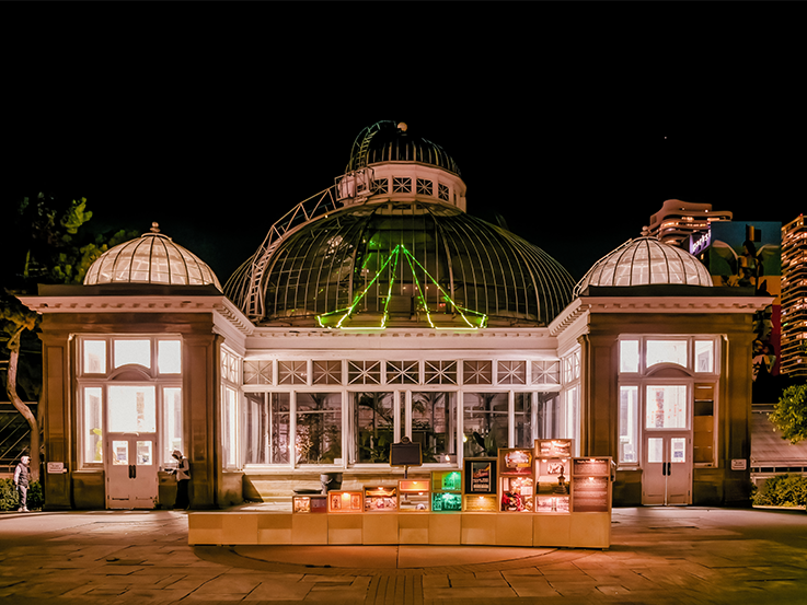 Allan Gardens Conservatory at night time with a projection of a tent on the glass dome. In front of it is a wooden art installation with green and white lights.